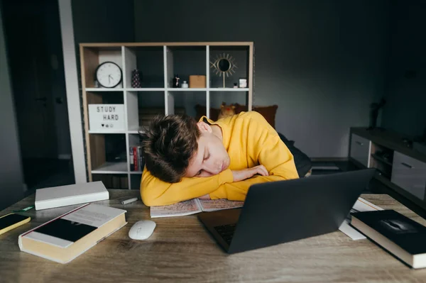 The student fell asleep at home studying on the desktop near books and laptops. Tired young man sleeping on the desk during online training. Quarantine.