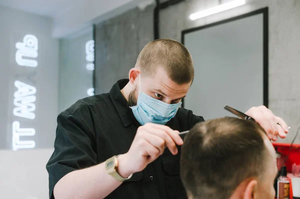 Professional barber in a medical mask cuts the client with scissors and a comb in a men\'s hairdresser with a concentrated face. Work of hairdressers in quarantine. coronavirus