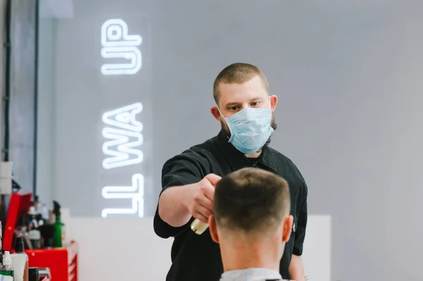 A professional hairdresser in a medical mask works in quarantine, cuts the client with a clipper. Portrait of a professional barber cuts a man in a medical mask. Coronavirus and services.