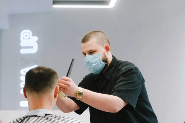 A barber man in a medical mask on his face cuts the client\'s hair in quarantine. Work of the service sector in quarantine. Hairdresser in a protective mask cuts the client.