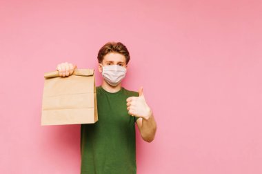 Tıbbi maskeli mutlu genç adam pembe arka planda, elinde kese kağıdıyla kameraya bakıyor, gülümsüyor ve baş parmağını kaldırıyor. Koronavirüs salgını. Karantinaya alın. covid-19.