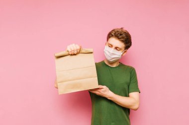 Günlük giysiler içinde ve tıbbi maske takmış yakışıklı bir genç elinde kese kağıdıyla pembe bir arka planda duruyor. Yemek teslimatı konsepti. Koronavirüs salgını. Karantinaya alın. covid-19