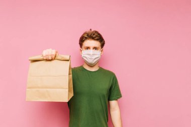 Koruyucu tıbbi maskeli yakışıklı bir genç pembe bir arka planda duruyor teslimattan bir paket yiyecekle, kameraya bakıyor ve gülümsüyor. Karantinaya alındı.