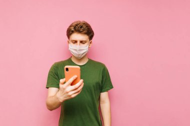 Tıbbi maskeli ve yeşil tişörtlü bir adam akıllı telefonunda internet kullanıyor ve koronavirüs haberlerini okuyor. Koronavirüs salgını. Karantinaya alın. Covid-19. Pembe arkaplan.