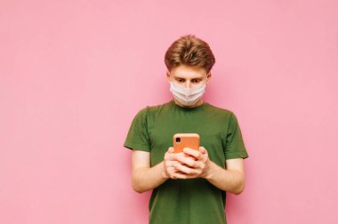 Tıbbi maskeli bir adam akıllı telefonuyla interneti kullanıyor ve koronavirüs haberlerini okuyor. Pembe arka plan. Koronavirüs salgını. Karantinaya alın. covid-19.