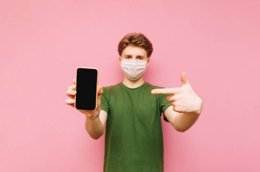 Tıbbi koruyucu maskeli genç adam pembe arka planda izole edilmiş, kameraya bakıyor, siyah ekranlı akıllı telefonu tutuyor ve parmağını işaret ediyor. Koronavirüs salgını. Karantinaya alın. covid-19.