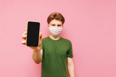 Yeşil tişörtlü ve koruyucu gazlı bez maskeli yakışıklı bir adam turuncu arka planda duruyor ve kameraya siyah ekranlı bir akıllı telefon gösteriyor. Koronavirüs salgını. Karantinaya alın. covid-19.