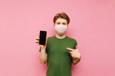 Gazlı bez maskeli yakışıklı bir adam pembe arka planda elinde akıllı bir telefonla duruyor, parmağını siyah ekrana doğrultuyor ve ciddi bir yüzle kameraya bakıyor..