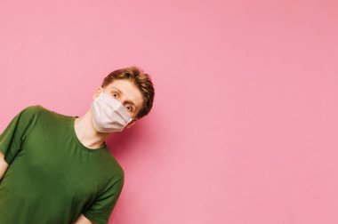 Yüzünde parlak günlük giysiler ve tıbbi maske olan komik bir adam yüzünde gülümsemeyle soldan bakıyor ve pembe arka plandaki kameraya bakıyor. Koronavirüs pandemiği. COVID-19