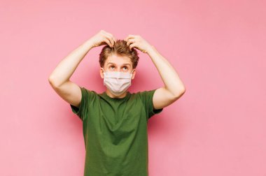 Tıbbi maskeli tatminsiz bir suratı olan tüylü sarışın bir adamın portresi pembe üzerine izole edilmiş, yeniden uzamış saçlarına dokunuyor. Adam karantinada saçının üstünü örtmüş. Barbreshop karantinası.