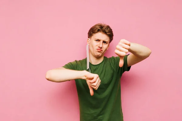 Upset young man in a mask removed from his face stands on a pink background, looks at camera and shows his finger down. Sad guy tired of wearing medical gauze masks he is dissatisfied.