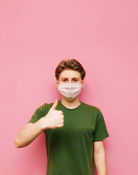 Portrait of positive guy in green t-shirt and medical mask on face isolated on pink background, looking at camera and showing thumbs up. Young man in a protective gauze mask shows a gesture like.