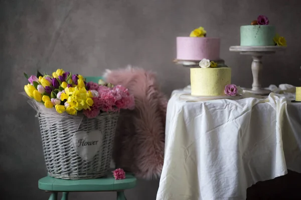 カラフルな春の花のバスケットは カラフルなイースター ケーキの近くに飾られた椅子の休日テーブルの上に配置 — ストック写真