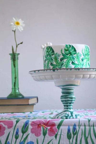 Holiday cake with palm leaves design. Tropical birthday party inspiration. Tasty cake with wafer paper monstera leaves on table with old books and flower in vase