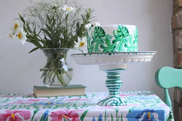 Holiday cake with palm leaves design. Tropical birthday party inspiration. Tasty cake with wafer paper monstera leaves on table with flowers in vase