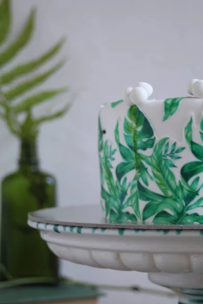 Holiday cake with palm leaves design. Tropical birthday party inspiration. Tasty cake with wafer paper monstera leaves on table with vase, flowers, old books and fern leaves