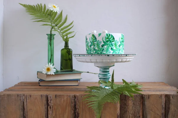 Holiday cake with palm leaves design. Tropical birthday party inspiration. Tasty cake with wafer paper monstera leaves on table with vase, flowers, old books and fern leaves