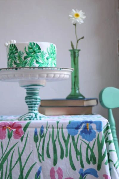 Holiday cake with palm leaves design. Tropical birthday party inspiration. Tasty cake with wafer paper monstera leaves on table with old books and flower in vase