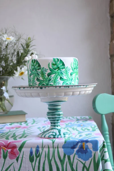 Holiday cake with palm leaves design. Tropical birthday party inspiration. Tasty cake with wafer paper monstera leaves on table with flowers in vase