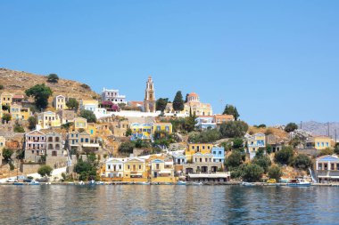 Symi, Yunanistan - 27 Haziran 2019: Suriye adasının çok renkli evleri, ağaçları ve halkları ve yaz günü tekneleri olan kıyı manzarası