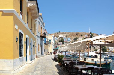 Symi, Yunanistan - 27 Haziran 2019: Suriye adasının çok renkli evleri, ağaçları ve halkları ve yaz günü tekneleri olan kıyı manzarası