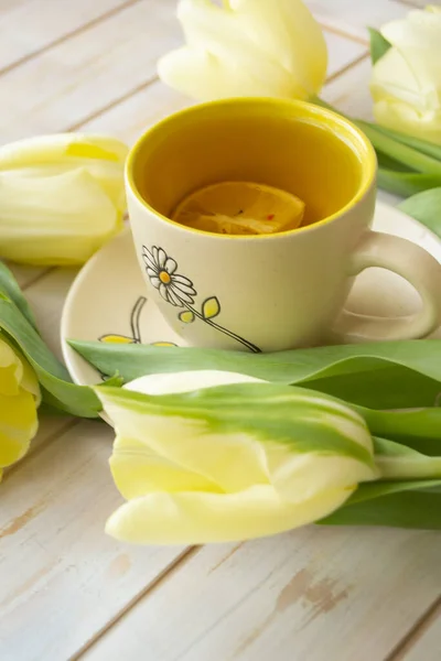 Black tea in the pastel yellow cup and lemon, yellow tulips on the white shabby wooden background. Vertical image — Stockfoto