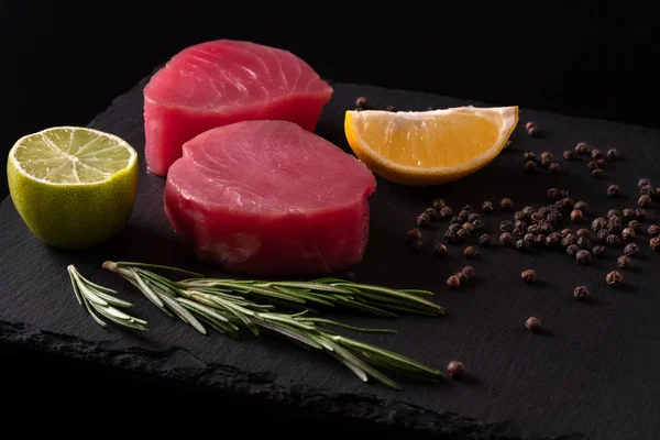 Fresh raw tuna fillet for steak on cutting board with lemon lime salt pepper and spices on dark black rustic background. Tasty fish dinner. Ingredients for cooking