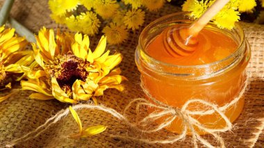 Bal kavanozu Tahta dallar cam kaseye yapışmış. Çiçek kokulu bal ile. Sağlıklı organik bal damlıyor, baldan yapılmış tahta kaşıktan dökülüyor. Çiçekli sarı arkaplan