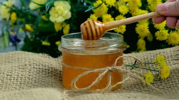 Honey jar. Dips wooden stick to glass bowl with liquid floral fresh honey. Healthy organic honey dripping, pouring from honey wooden spoon. Yellow background with flowers