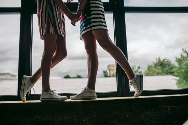 两个穿着连衣裙和运动鞋的女孩站在大窗户边牵着手 — 图库照片