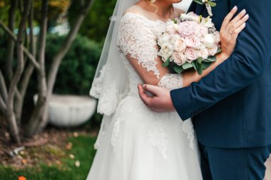 Damat parkta gelini kucaklıyor, gelin elinde bir buket pembe çiçek tutuyor.