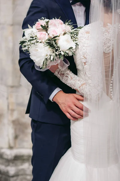 黒のスーツの新郎は花嫁を白いウェディングドレスとベールで抱きしめる 花嫁は花束を持ってる — ストック写真