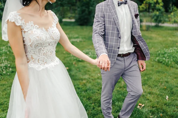 灰色のスーツの新郎と結婚式のドレスとベールの花嫁は緑の芝生の上を歩いています — ストック写真