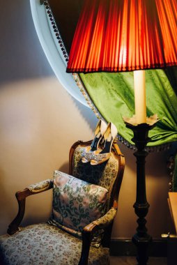 Düğün yüksek topuklu ayakkabıları güzel bir sandalyenin arkasında yatıyor, sandalye yeşil perdeli bir pencerenin altında duruyor, yanında kırmızı bir lamba var.