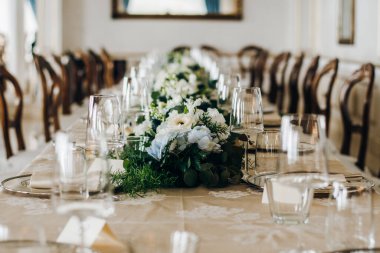 Düğün masası çiçek aranjmanları, bardaklar, tabaklar ve çatal bıçaklarla süslenmiştir. Masa güzel bir masa örtüsüyle kaplıdır.