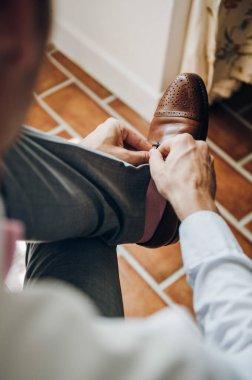 Damat bir sandalyede oturuyor ve ayakkabı bağcıklarını kahverengi ayakkabılara bağlıyor.
