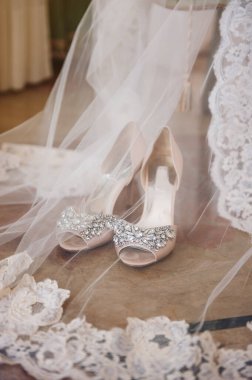 Şık gelinin yüksek topuklu ayakkabıları, taşlarla süslenmiş, beyaz dantelli bir peçenin yanında yerde duruyor.