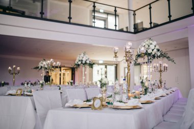 Düğün şölen alanında yeşil masa örtüleri olan masalar var. Masaların üzerinde çiçekler, yeşiller, mumlar, çatal bıçaklar, tabaklar ve peçeteler var. İki şişe şampanya var.