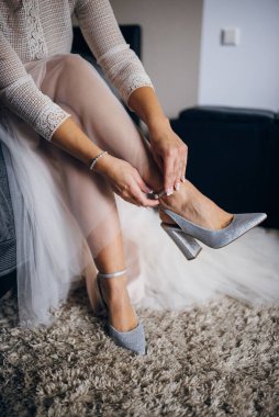 Gelin, beyaz bir gelinlik içinde bir kanepeye oturur ve gümüş topuklu ayakkabılar giyer.