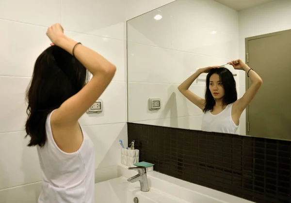 Mulher asiática escovando o cabelo na frente do espelho no banheiro — Fotografia de Stock