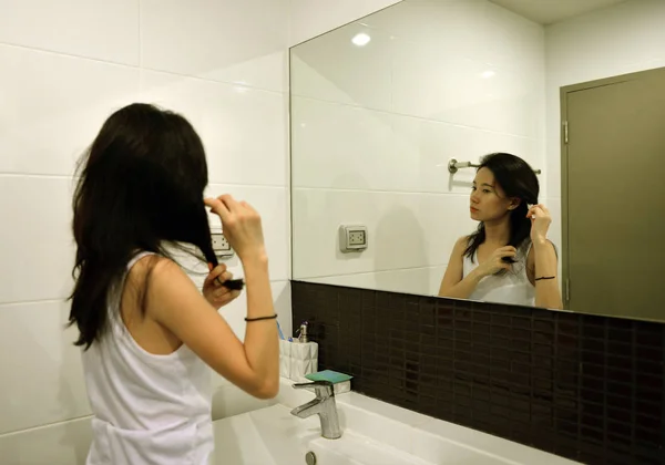 Mulher asiática escovando o cabelo na frente do espelho no banheiro — Fotografia de Stock