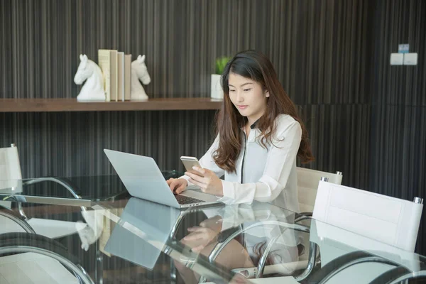 An Asian business woman using a phone in front of a laptop — Stock Photo, Image