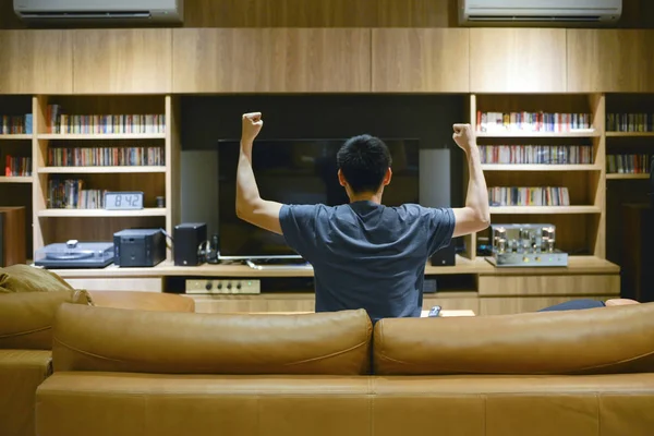 Asian man cheering in front of TV in living room at night