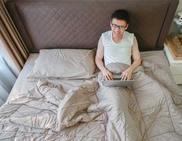 Homem asiático feliz usando um laptop na cama no quarto — Fotografia de Stock