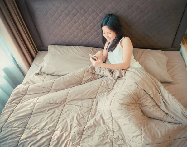 Felice donna asiatica utilizzando uno smartphone sul letto in camera da letto — Foto Stock