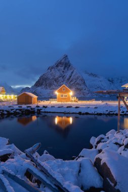 Geceleri ev, kulübe ya da ev, Norveç 'in Reine City, Lofoten adalarındaki balıkçı köyü, Nordland County, Norveç, Avrupa. Beyaz karlı dağ tepeleri, kış mevsiminde doğa manzarası.