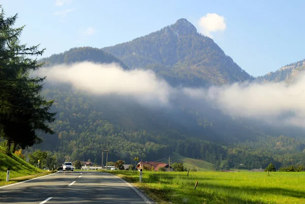 Дорога между зелеными лугами с горами в облаках на заднем плане в солнечный день . — стоковое фото