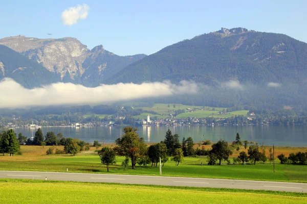 El camino entre verdes prados con las montañas y un lago en las nubes en el fondo en el día soleado . —  Fotos de Stock