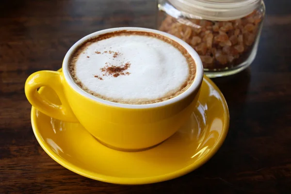 Cangkir kuning cappuccino dalam cangkir kuning dan botol kaca gula di latar belakang kayu . — Stok Foto