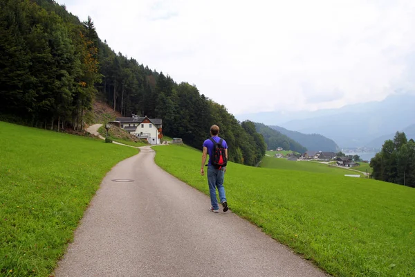 Podróż do Sankt-Wolfgang, Austria. Młody człowiek chodzą na drodze między polami z domów i gór na tle. — Zdjęcie stockowe